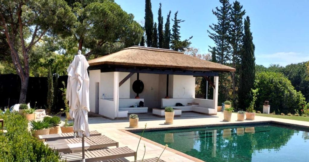 A contemporary cape reed thatched gazebo standing on concrete pillars, surrounded by lush greenery.