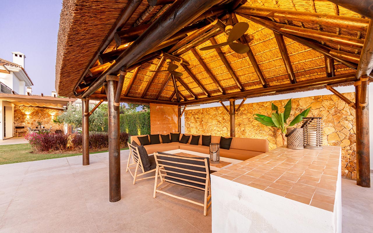 Zona de entretenimiento exterior con gazebo de junco africano, sofá de obra, barra de terracota y muro de piedra en Marbella